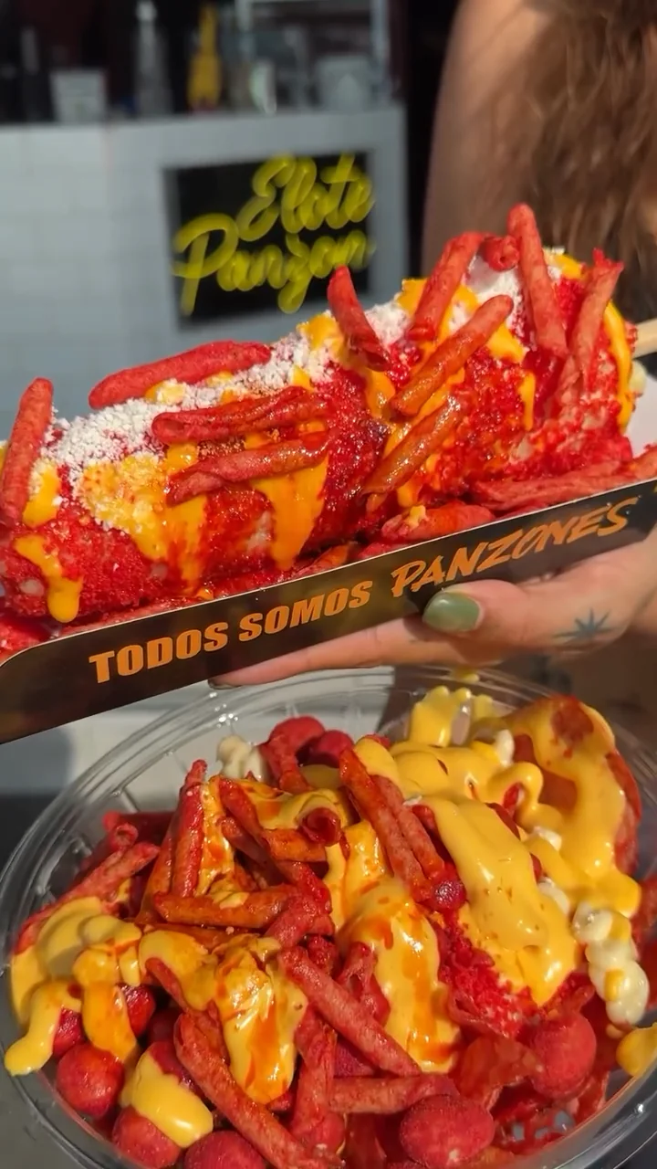 Elote cubierto de Takis y queso cheddar en su charola «Todos somos panzones», sobre un tazón con Takis y queso
